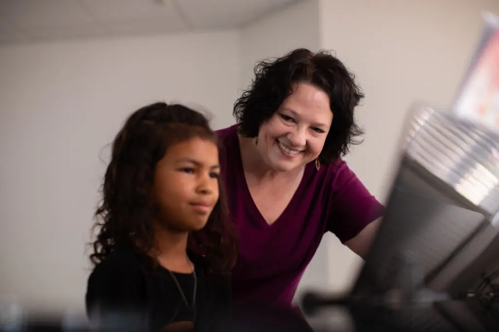 Music teacher Amy Rau leans over smiling at a child playing the piano at Cavod Performing Arts | Private Music Lessons at Cavod | Cavod Performing Arts