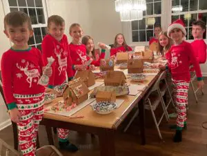 Children in matching Christmas pajamas smiling while making ginger bread houses.