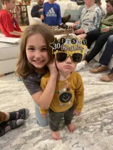 Two children smiling while one comically wears a set of sunglasses that say "Its my 30th birthday".