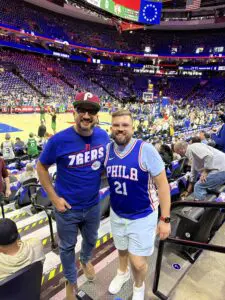 Two men smiling and posing for a picture at a Philadelphia 76ers game.