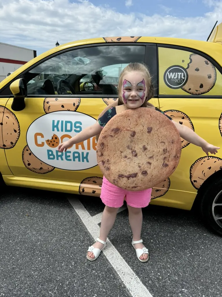A small child dressed as a cookie behind a car designed around the company :"Kids Cookie Break"