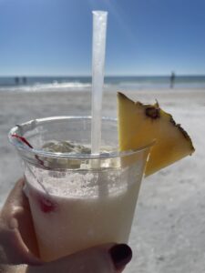 A close up of a tropical drink with a pineapple wedge with the beach on a sunny day in the background.